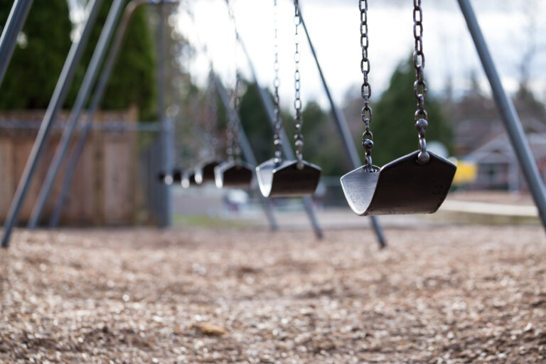 Eine Reihe leerer Schaukeln, die an Ketten auf einem Spielplatz hängen. Die Schaukelsitze sind schwarz und in gleichmäßigen Abständen angeordnet, mit einem Hintergrund aus Holzspänen auf dem Boden und einer verschwommenen Sicht auf Bäume und einen Zaun in der Ferne. Das Bild scheint an einem bewölkten Tag aufgenommen worden zu sein.